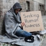 Homeless man showing "Seeking Human Kindness" sign on cardboard